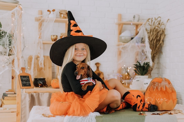 Une petite fille blonde dans un costume de sorcière avec une jupe bouffante orange tient un teckel nain Halloween