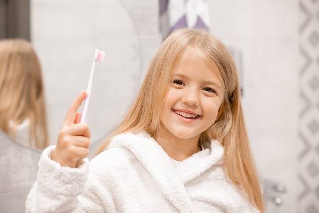 petite fille blonde dans un peignoir blanc se brosse les dents devant le miroir de la salle de bain