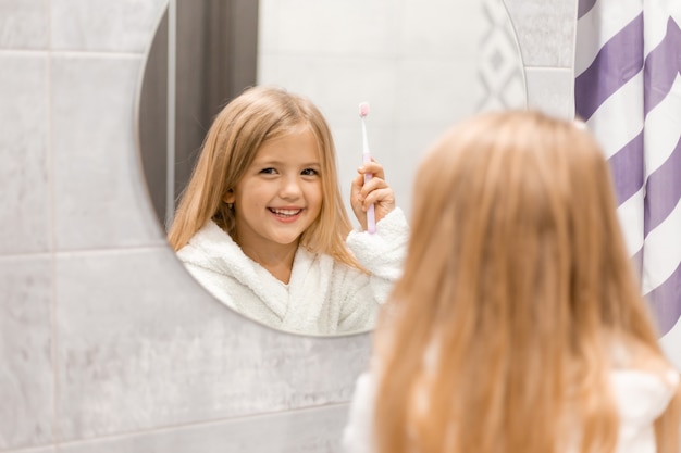 petite fille blonde dans un peignoir blanc se brosse les dents devant le miroir de la salle de bain