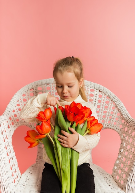 Petite fille blonde dans un pull blanc est assis sur une chaise blanche et regarde un bouquet de tulipes rouges sur un mur rose