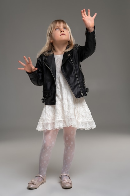 La petite fille blonde dans une robe blanche et une veste en cuir noire pose