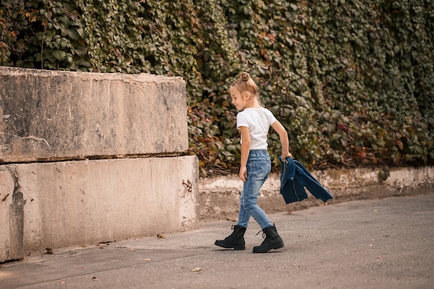 Une petite fille blonde élégante en jeans et un T-shirt blanc marche dans la rue. Fille 7 ans petit modèle, bel enfant