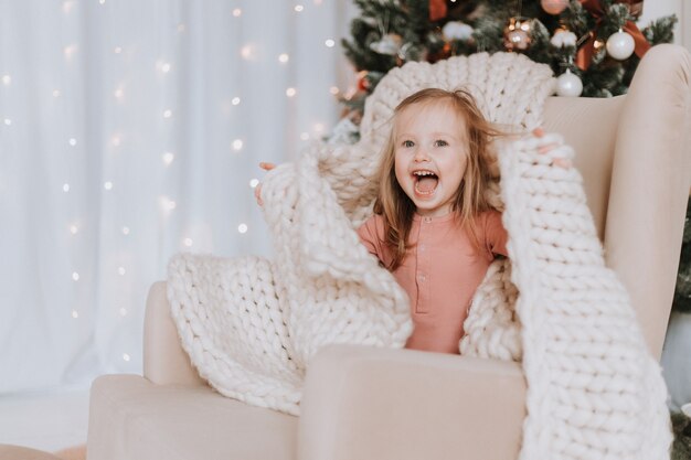 petite fille blonde enveloppée dans un grand plaid tricoté est assise sur une chaise à côté de l'arbre de Noël