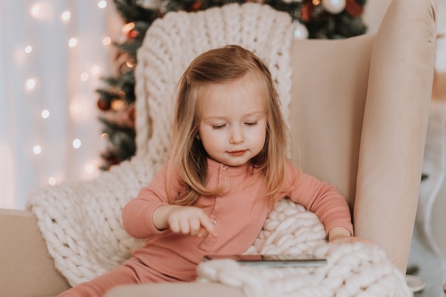 petite fille blonde enveloppée dans un grand plaid tricoté est assise sur une chaise à côté de l'arbre de Noël