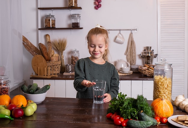 Une petite fille blonde est assise à la table de la cuisine et boit des vitamines