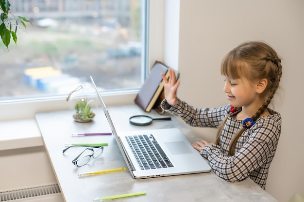 Petite fille blonde à faire ses devoirs à la maison à la table. L'enfant est scolarisé à la maison. Une fille aux cheveux clairs effectue une tâche en ligne à l'aide d'un ordinateur portable et d'une tablette.