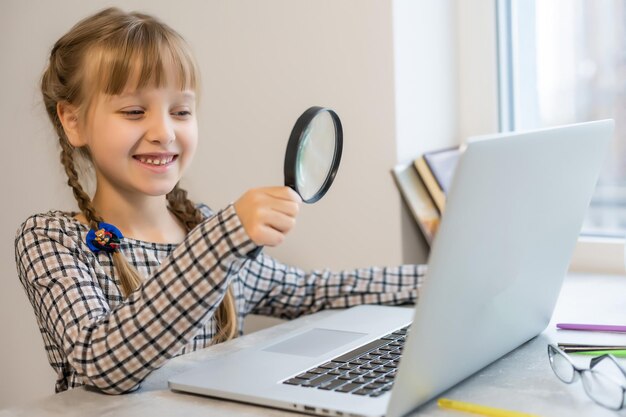 Petite fille blonde à faire ses devoirs à la maison à la table. L'enfant est scolarisé à la maison. Une fille aux cheveux clairs effectue une tâche en ligne à l'aide d'un ordinateur portable et d'une tablette.