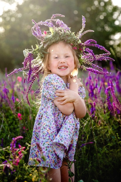 Une petite fille blonde heureuse dans une couronne de sauge et une robe colorée dans un champ fleuri d'été