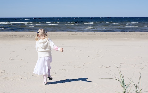 Petite fille blonde heureuse en robe blanche se promène sur la plage sur la côte de la mer Baltique.