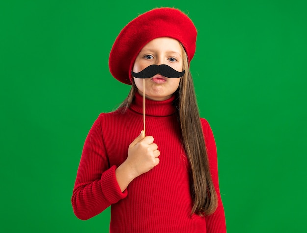 Petite fille blonde ludique portant un béret rouge essayant une fausse moustache regardant l'avant isolé sur un mur vert avec espace pour copie