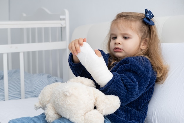 Petite fille blonde avec la main en plâtre assis dans son lit.