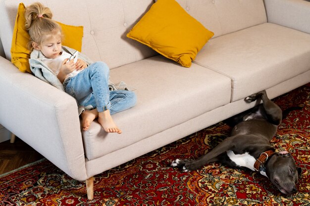 Une petite fille blonde à la maison sur le canapé avec un téléphone portable et un gros chien, des enfants et des gadgets.