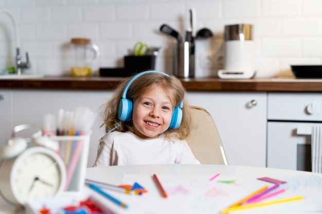 Petite fille blonde à la maison dans la cuisine à table avec des écouteurs, dessine et apprend à distance.
