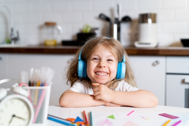 Petite fille blonde à la maison dans la cuisine à table avec des écouteurs, dessine et apprend à distance.