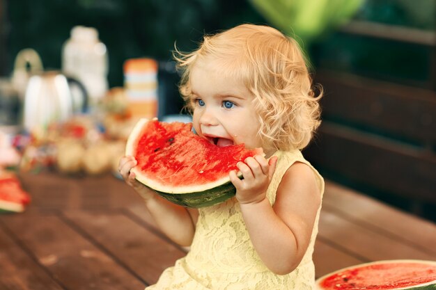 Petite Fille Blonde Mange Une Pastèque Juteuse Dans Le Jardin. Les Enfants Mangent Des Fruits Dans La Rue. Une Alimentation Saine Pour Les Enfants. Jardinage Pour Tout-petits.