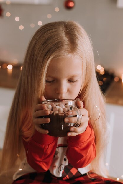 Petite fille blonde en pyjama de noël rouge boit du cacao avec des guimauves dans une cuisine décorée. maison confortable les week-ends d'hiver et les vacances. mode de vie. espace pour le texte. Photo de haute qualité