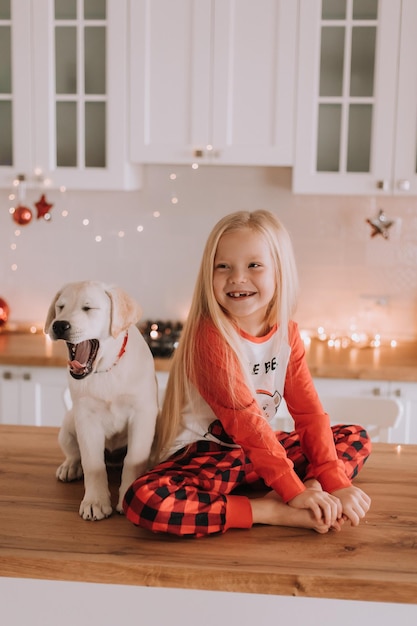 Une petite fille blonde en pyjama de Noël rouge est assise dans une cuisine ornée de guirlandes avec un chien blanc. week-ends et vacances d'hiver dans un cercle familial chaleureux. enfant et animal de compagnie. espace pour le texte. Photo de haute qualité