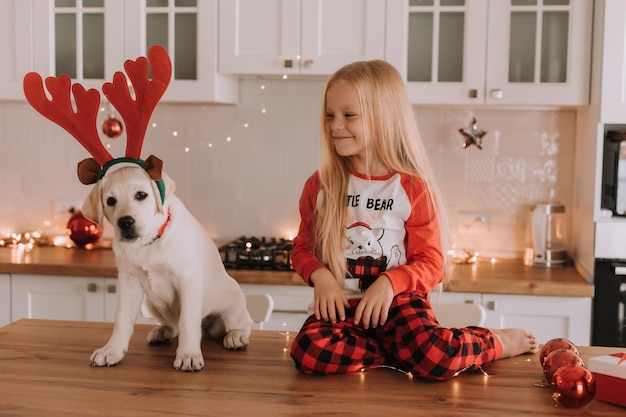 Une petite fille blonde en pyjama de Noël rouge met des cornes de cerf sur la tête d'un chien. intérieur festif. l'enfant joue avec son animal de compagnie bien-aimé. mode de vie. espace pour le texte. Photo de haute qualité