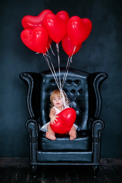 Petite fille blonde en robe blanche avec ruban rouge assis sur le fauteuil avec ballon coeur rouge le jour de la Saint-Valentin