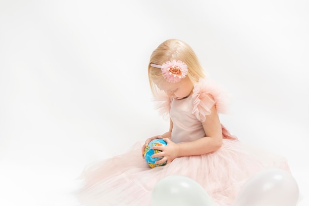 Petite fille blonde en robe rose est assise sur fond blanc et regarde un petit globe dans les mains Vacances pour enfants