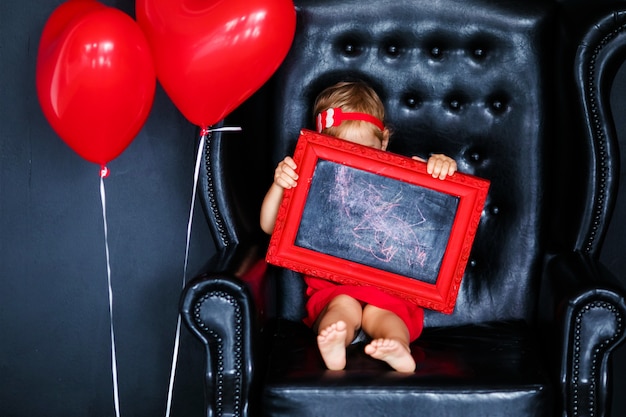 Petite fille blonde en robe rouge avec une couronne rouge avec des coeurs, assise sur le fauteuil avec ballon coeur rouge le jour de la Saint-Valentin.