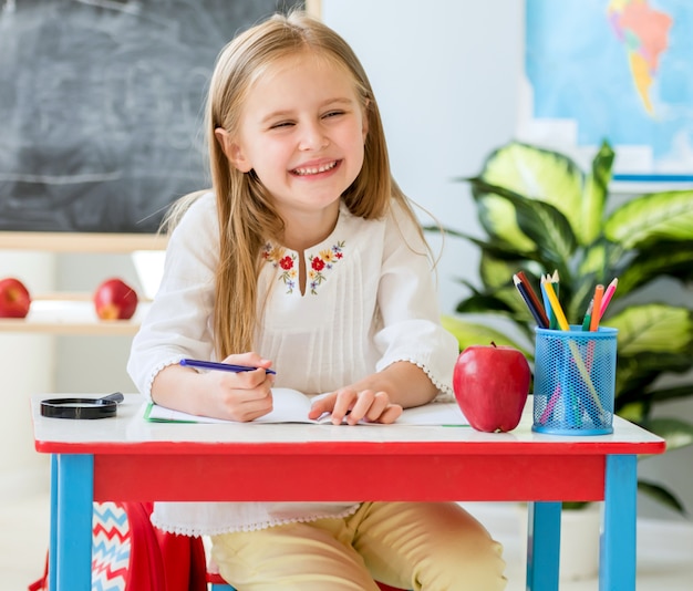 Petite fille blonde s'asseoir au bureau blanc et rire dans la salle de classe