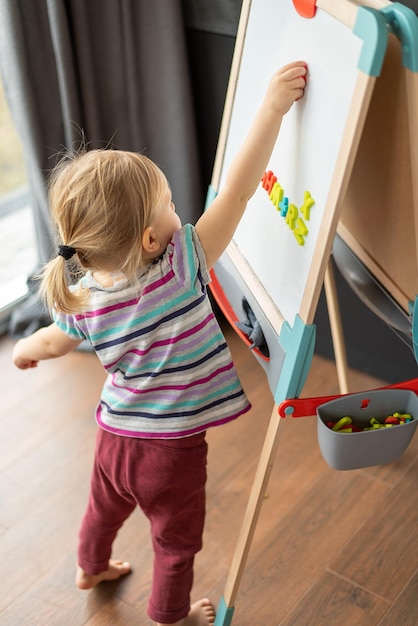 Photo une petite fille blonde se tient près d'un tableau magnétique sur un chevalet pour enfants dans la chambre