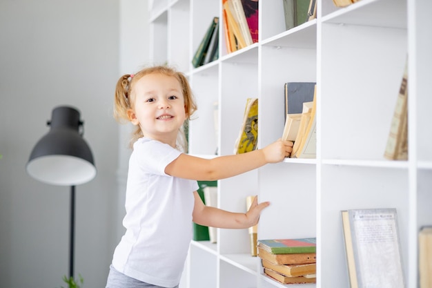 Une petite fille blonde sort un livre dans une bibliothèque lumineuse à la maison pour enseigner et lire des livres pour enfants