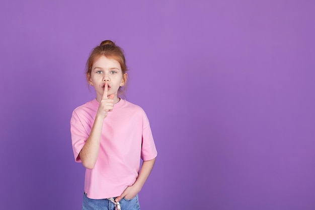 Une petite fille blonde souriante et joyeuse tient ses bras croisés pliés isolément sur un fond violet portrait en studio pour enfants Le concept de mode de vie dans l'enfance