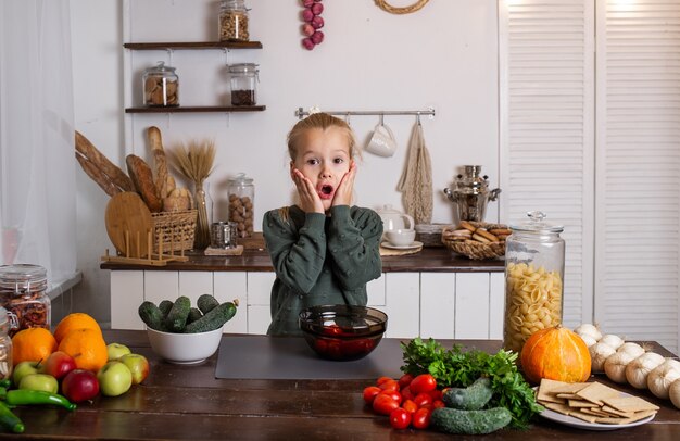 Une petite fille blonde surprise est assise à une table dans la cuisine avec des légumes et des fruits