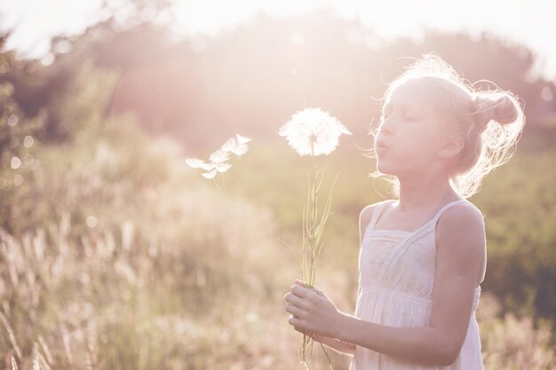 Photo petite fille blonde tenant un pissenlit et le soufflant pendant les vacances d'été et l'humeur au coucher du soleil