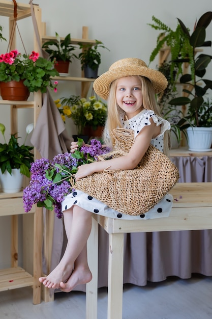 petite fille blonde vêtue d'une robe à pois noirs et d'un chapeau de paille avec un bouquet de fleurs lilas