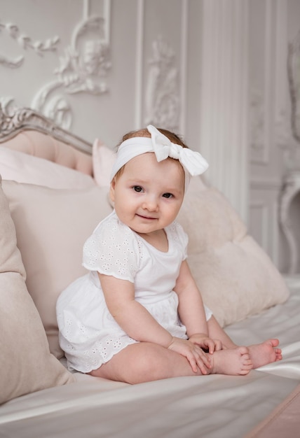 Une petite fille en body blanc et avec un bandage sur la tête est assise sur le lit dans la chambre