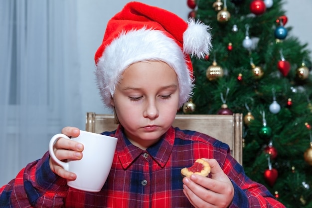 La petite fille boit du thé et des biscuits à l'arrière-plan de l'arbre de Noël