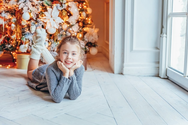 Petite fille avec boîte-cadeau près de l'arbre de Noël la veille de Noël à la maison. Jeune enfant dans une chambre lumineuse avec décoration d'hiver Famille heureuse à la maison. Noël Nouvel An décembre pour le concept de célébration