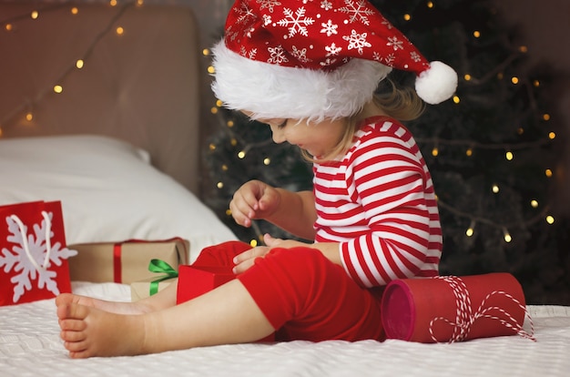 Petite fille en bonnet de Noel ouvrant la boîte-cadeau de Noël Enfant en bas âge drôle et expressif appréciant des cadeaux