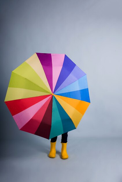 Une petite fille en bottes de caoutchouc jaune s'est couverte d'un parapluie multicolore sur fond bleu avec une copie de l'espace