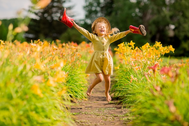 petite fille en bottes de caoutchouc rouges et un chapeau de paille arrosant des fleurs rouges dans le jardin