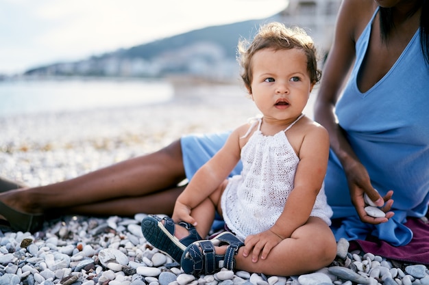 Petite fille avec une bouche ouverte est assise sur la plage près de sa mère tenant des cailloux dans sa main