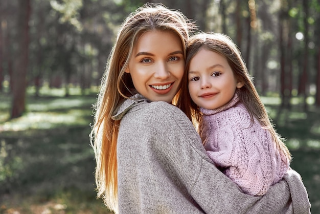 Une petite fille bouclée en pull embrasse sa mère dans les bois. Saison froide, le soleil éclatant est vu à travers les arbres. Elle porte un pull rose et la jeune femme est en pull gris