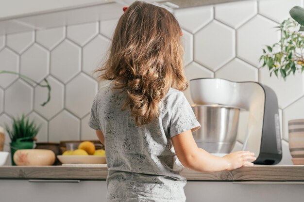 Petite fille brune de 3 ans fait une tarte aux pommes dans la cuisine. L'enfant se tient près du mélangeur planétaire. Vue arrière