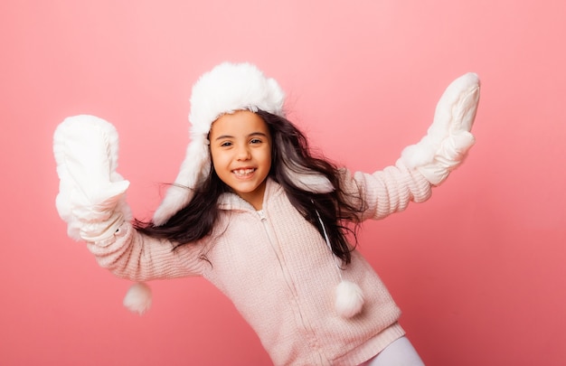 Petite fille brune aux cheveux longs dans un chapeau d'hiver et des mitaines sur fond rose