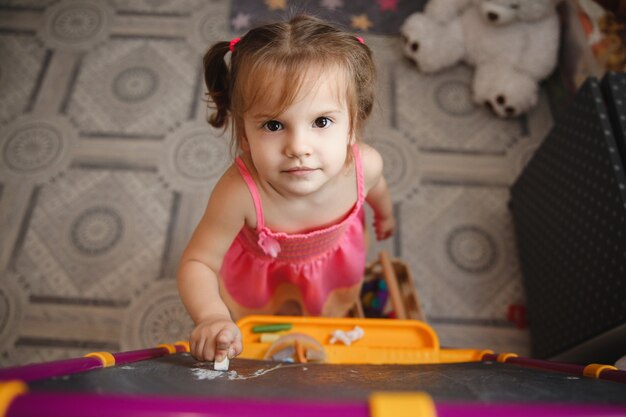 Une petite fille brune avec deux queues de cheval à la maison dans la salle de jeux près du tableau noir avec de la craie dans ses mains.