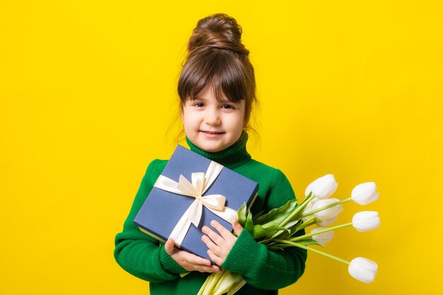 Une petite fille brune heureuse tient une boîte-cadeau et un bouquet de tulipes sur fond jaune dans le studio Le concept de la fête des mères le 8 mars Saint-Valentin