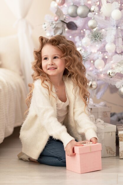 Petite fille avec un cadeau rose, présente sous le sapin de Noël blanc. Bonne enfance et vacances à la maison.