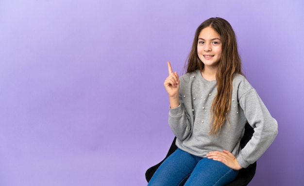 Petite fille caucasienne assise sur une chaise isolée sur fond violet montrant et levant un doigt en signe du meilleur