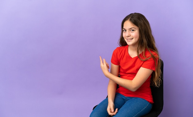 Petite fille caucasienne assise sur une chaise isolée sur fond violet pointant vers l'arrière