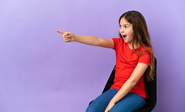 Petite fille caucasienne assise sur une chaise isolée sur fond violet pointant vers l'extérieur