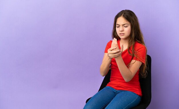 Petite fille caucasienne assise sur une chaise isolée sur fond violet souffrant de douleurs dans les mains