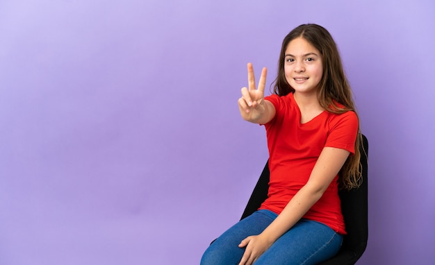 Petite fille caucasienne assise sur une chaise isolée sur fond violet souriant et montrant le signe de la victoire